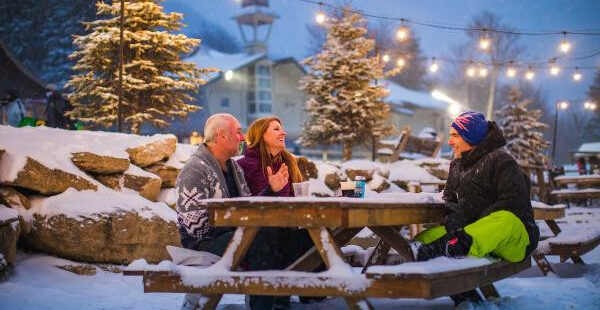 Three people enjoy an outdoor picnic on a lightly snow-covered table, surrounded by snow-dusted trees and string lights.