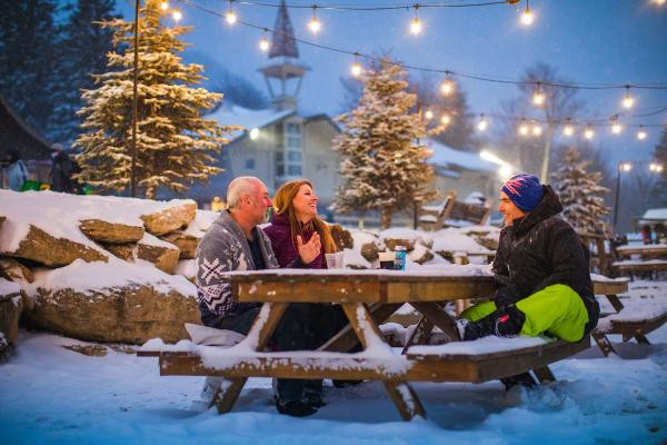 Three people enjoy an outdoor picnic on a lightly snow-covered table, surrounded by snow-dusted trees and string lights.
