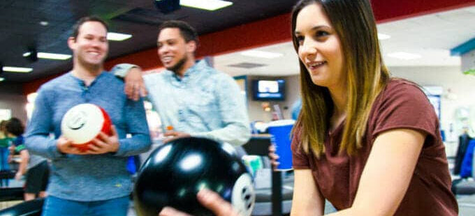 Hình ảnh một cô gái trẻ đang chơi bowling với hai người bạn đứng phía sau theo dõi và mỉm cười, thể hiện không khí vui vẻ tại khu bowling của Triple Play Resort Idaho