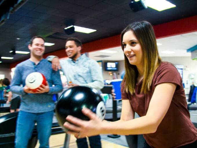 Hình ảnh một cô gái trẻ đang chơi bowling với hai người bạn đứng phía sau theo dõi và mỉm cười, thể hiện không khí vui vẻ tại khu bowling của Triple Play Resort Idaho